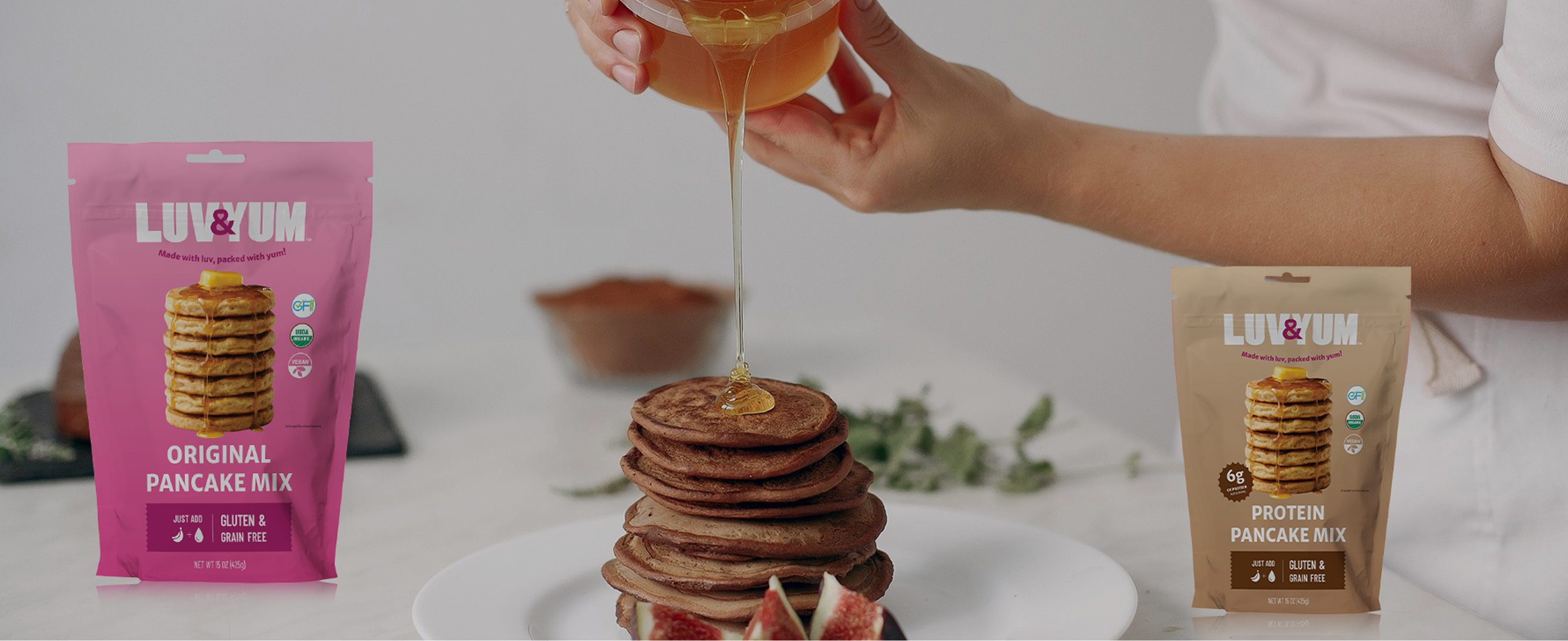 Person pouring syrup onto a stack of pancakes with LUVYUM pancake mix packages in the foreground.